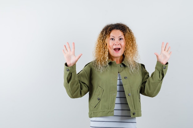 Woman with curly blonde hair showing palms in surrender gesture in green jacket and looking wondered , front view.