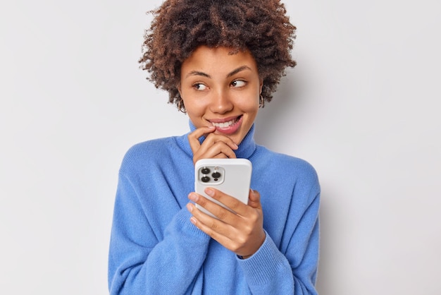 woman with curly hair uses smartphone applicatio looks with smile away enjoys shopping discounts communicates with friend online dressed casually poses against white