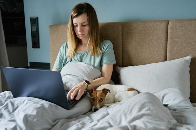 Woman with cute dog lying in bed and using laptop at morning