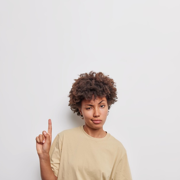 woman with dark curly bushy hair indicates above showscopy space suggests going upstairs wears casual beige t shirt isolated on white