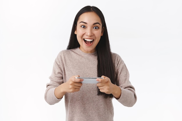 woman with dark long hair, holding smartphone horizontally, achieve success, win game, look camera impressed