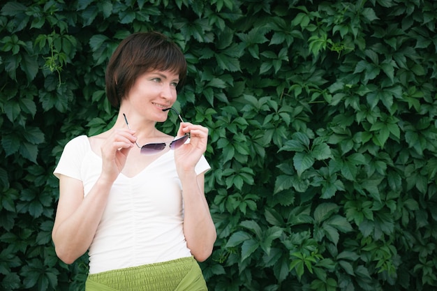 Woman with dark red hair stands and holds sunglasses in her hands n front of a wall covered in leaves