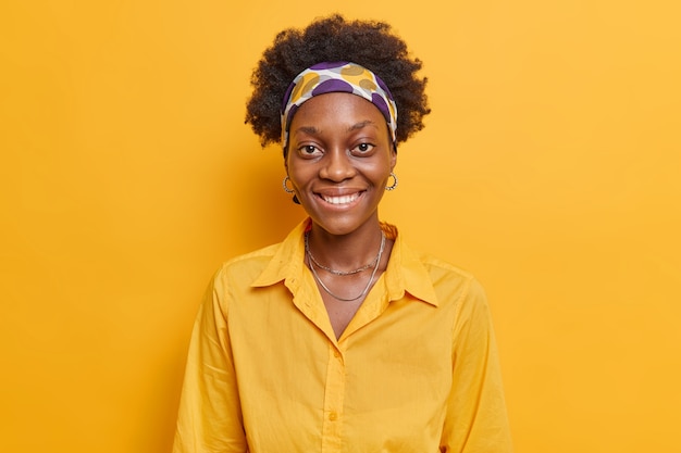  woman with dark skin smiles positively happy to get compliment expresses cheerful emotions comes on informal meeting wears casual shirt isolated on yellow wall