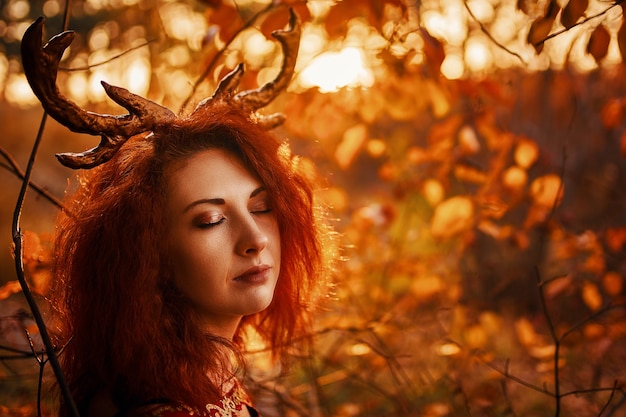 Woman with deer horns in autumn forest beautiful mystery gothic creature in long red cloak with deer...