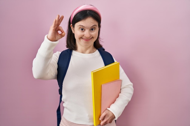 Woman with down syndrome wearing student backpack and holding books smiling positive doing ok sign with hand and fingers successful expression