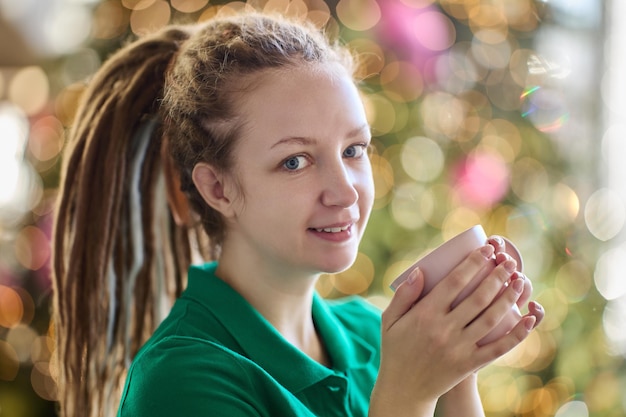 Woman with dreadlocks is holding cup of coffee with Christmas tree on background.