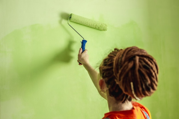 Woman with dreadlocks in an orange clothes is painting the wall with a roller in green color Concept of repair renovation of the new apartment Copy space and closeup