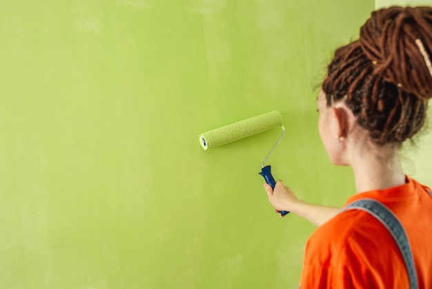 Woman with dreadlocks in an orange tshirt and denim jumpsuit is painting the wall with a roller in green color Concept of repair renovation of the new apartment Copy space