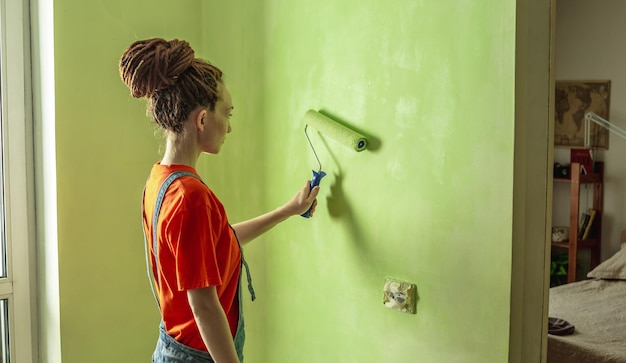 Woman with dreadlocks in an orange tshirt and denim jumpsuit is painting the wall with a roller in green color Concept of repair renovation of the new apartment Copy space