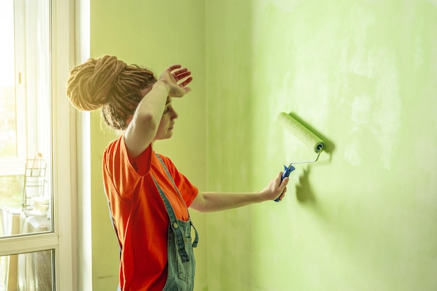 Woman with dreadlocks in an orange tshirt and denim jumpsuit is painting the wall with a roller in green color Concept of repair renovation of the new apartment