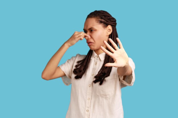 Woman with dreadlocks pinching nose stop breathing bad odor disgusted by smell of farting her grimace expressing repulsion showing stop gesture Indoor studio shot isolated on blue background