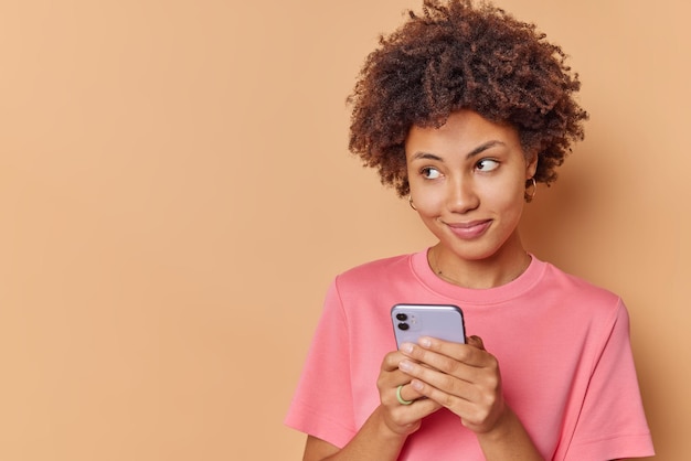 woman with dreamy expression uses mobile phone sends text messages to friend dressed casually concentrated away poses against beige blank copy space on left