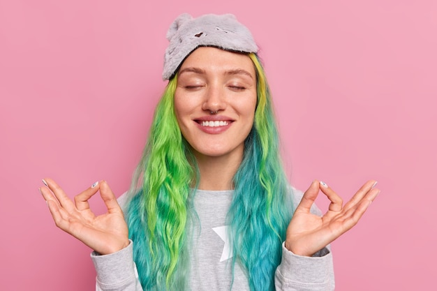  woman with dyed colorful hair keeps eyes closed makes mudra gesture relaxes after studying or work meditates before sleep wears nightwear poses on pink 