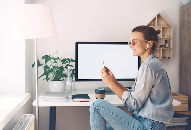 Woman with earphones wearing glasses using smartphone while working on computer from home Female entrepreneur small business responding on emails or calls from clients Modern creative workplace