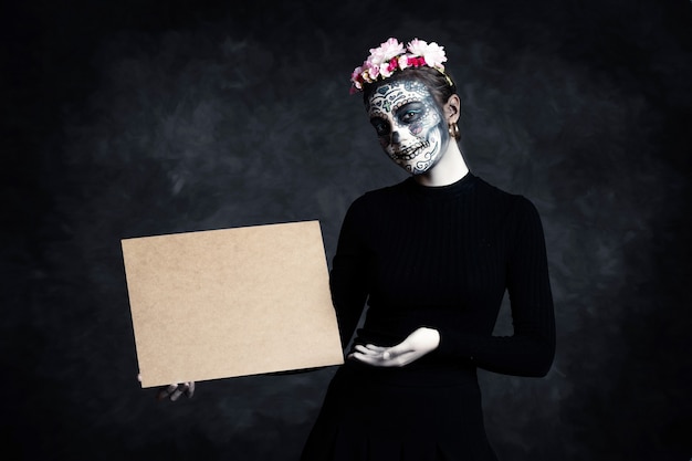 Woman with face painted as a Catrina and flowers in her hair on dark background pointing at poster. Day of the Dead