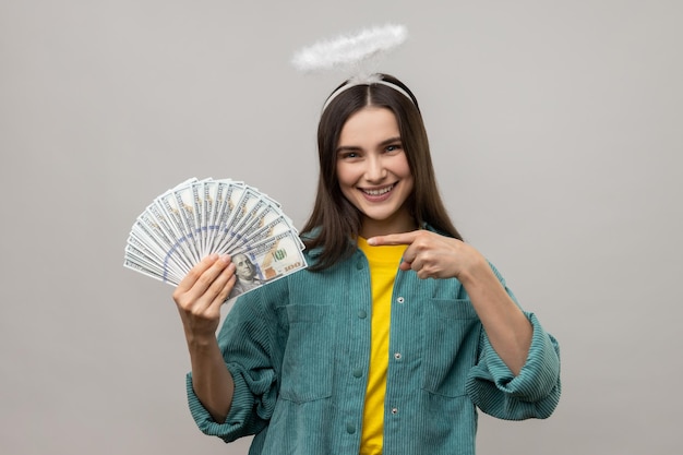 Woman with halo on head pointing finger on bunch of dollar banknotes and smiling profit