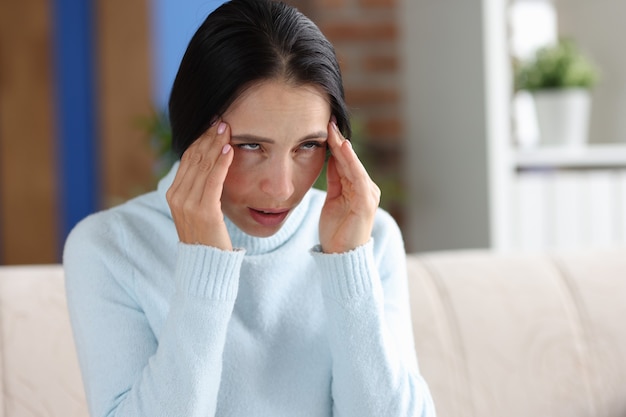 Woman with headache holds hands at temples while sitting on couch. Head migraine symptoms