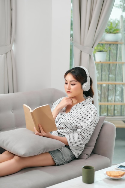 Woman with headphones reading a book in the living room sitting on the couch