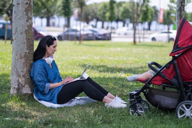 woman with headphones working on tablet computer while baby sleeping in buggy on summer park lawn