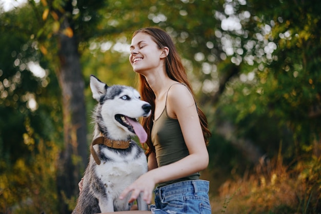 A woman with a husky breed dog smiles and affectionately strokes her beloved dog while walking in nature in the park in autumn against the backdrop of sunset