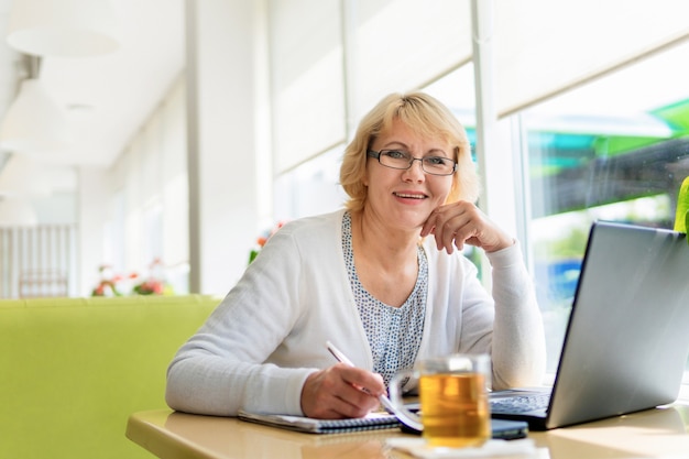 A woman with a laptop is working in an office. A middle-aged woman is a businesswoman in a cafe. She smiles and writes in her notebook.