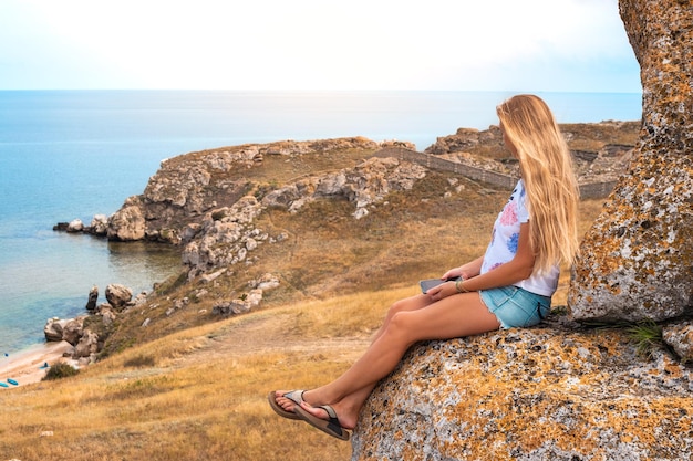 A woman with long blond hair sits on a rock on a mountain overlooking a rocky seashore Travel and tourism