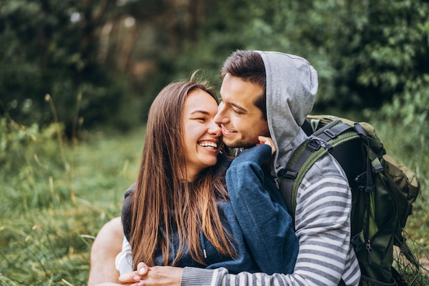 Woman with long hair and man sitting on the grass in the forest with backpacks. Have fun in nature, hugging and kissing