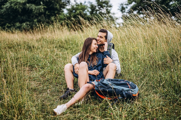 Woman with long hair and man sitting on the grass in the forest with backpacks. Have fun in nature, hugging and kissing