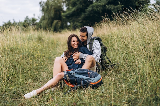 Woman with long hair and man sitting on the grass in the forest with backpacks. Have fun in nature, hugging and kissing