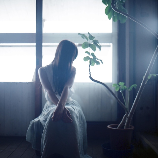 Woman with long hair sitting by potted plant against window