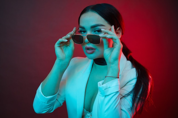 Woman with long hair and sunglasses in a white shirt poses in neon light in the studio.