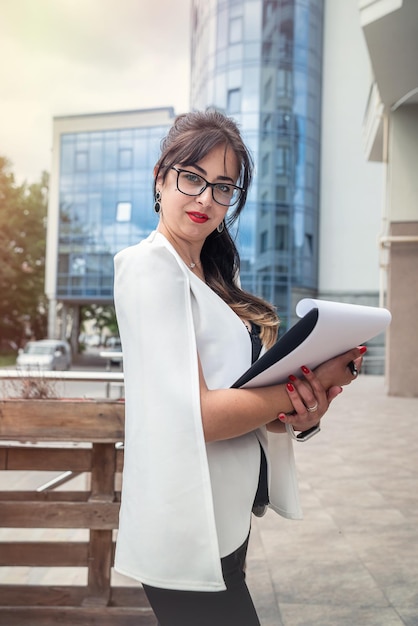 A woman with luxurious hair in glasses with documents walks through a beautiful city