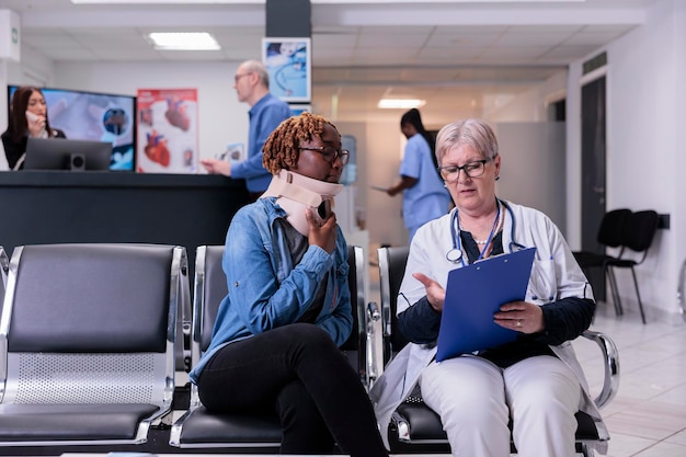 Woman with medical neck collar at examination with doctor, sitting in waiting room lobby. Patient with cervical foam and senior medic filling in checkup report papers at appointment in hospital.