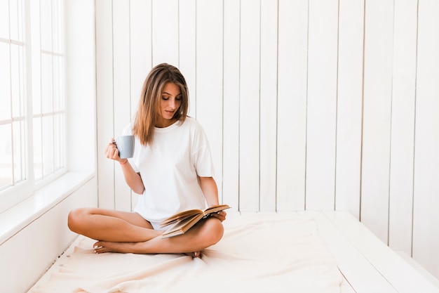 Woman with mug reading book on white surface