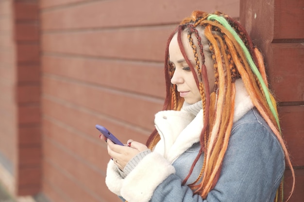 Woman with orange dreadlocks and in a warm denim jacket using smartphone on red wooden background