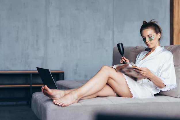 Woman with patches under her eyes sits on the couch and reads a magazine