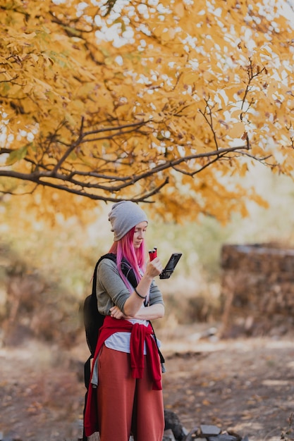 A woman with pink hair near a tree with yellow leaves holds a phone with a mechanical stabilizer