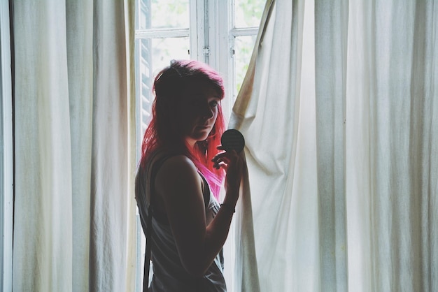 Woman with pink hair standing by curtain against window at home