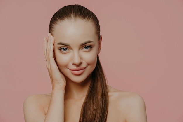 Woman with ponytail enjoys fresh skin charming look bare shoulders isolated on pink wall Health care makeup spa