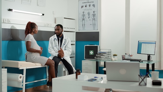 Woman with pregnancy belly talking to doctor at examination in office, meeting to discuss about childbirth. Future mother attending consultation appointment with general practitioner.