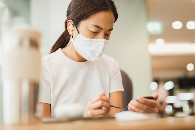 Woman with protective mask working on smartphone with wireless earphones in cafe.