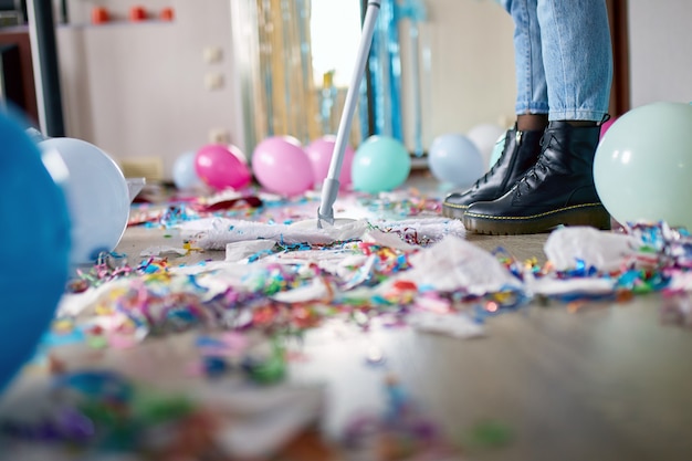 Woman with pushbroom cleaning mess of floor in room after party confetti, morning after party celebration, housework, cleaning service