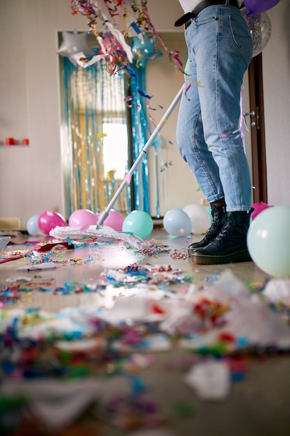 Woman with pushbroom cleaning mess of floor in room after party confetti