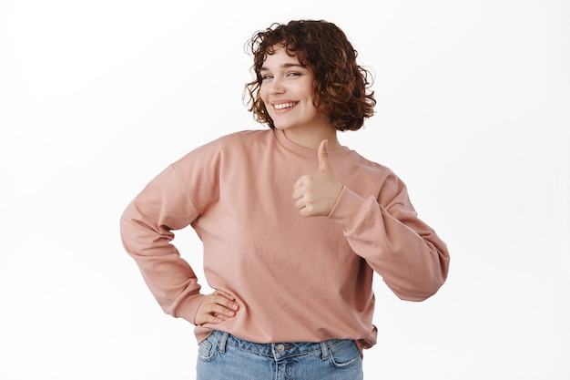 woman with short curly hairstyle, shows yes thumb up gesture and smiles pleased, satisfied by something good, give approval, agree and like, standing on white