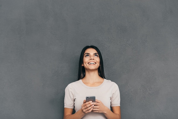 Woman with smart phone. Attractive young woman holding smart phone and looking up with smile while standing against grey background