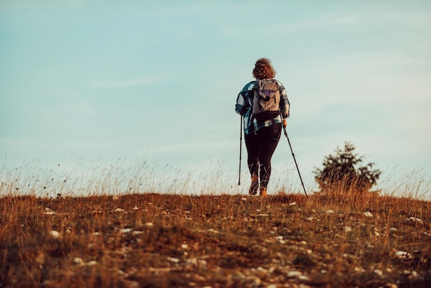 A woman with sticks a backpack on her back and mountaineering equipment walking on top of a mountain at sunset