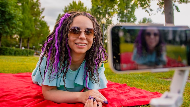 A woman with a stylish hairstyle lies on a blanket in the park and records a video blog using her smartphone A female blogger shoots a video on a camera on a tripod on a sunny summer day