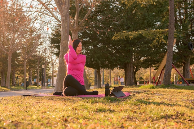 Woman with a tablet performing yoga exercises in a park at sunset