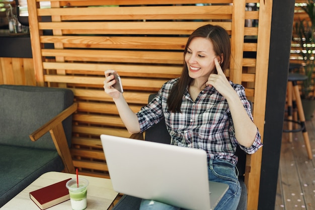 Woman in wooden outdoors street summer coffee shop sitting with laptop pc computer, doing selfie shot on mobile phone