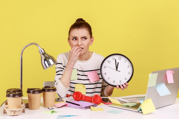 Woman worker biting nails holding big wall clock sitting at workplace all covered with sticky notes